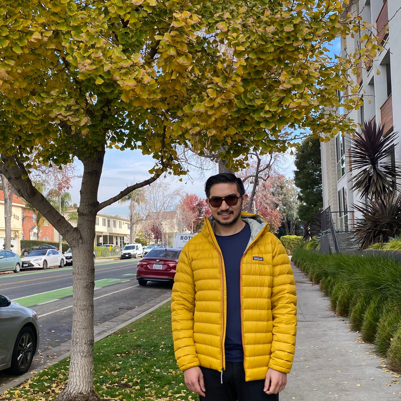 Person wearing a yellow puffer jacket standing on a sidewalk with trees and cars in the background